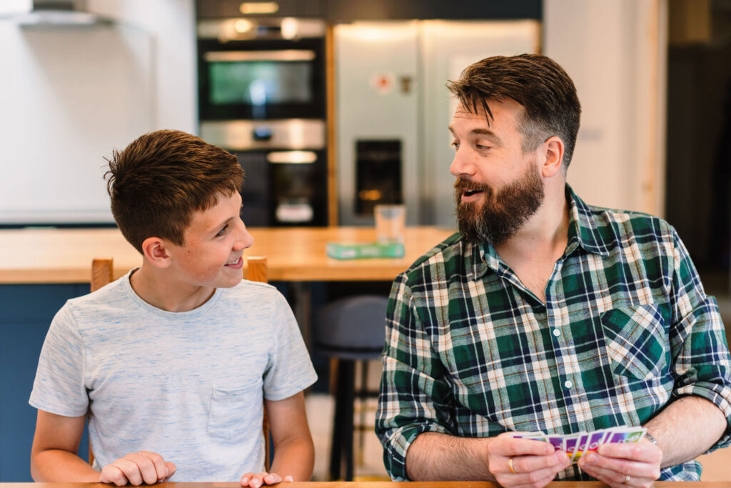 Smiling father and son sitting at a table, talking and playing a card game together.